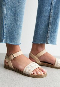 Sandales beiges avec un design supérieur tressé, sangle de cheville ajustable avec boucle en métal, et semelle en caoutchouc plate, associées à un jean cropped bleu clair.
