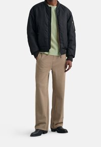 Giacca bomber nera, maglione verde a maglia, pantaloni beige a motivo houndstooth, scarpe in pelle nere, vestibilità rilassata, texture delicata sui tessuti.