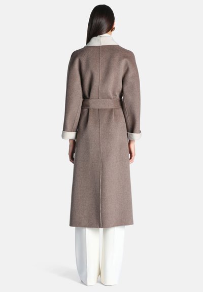 Femme aux longs cheveux foncés portant un long manteau marron ceinturé avec des poignets et un col blancs, assorti à un pantalon blanc large, vue de dos.