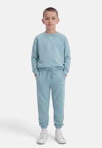 Αγόρι στέκεται με τα χέρια στις τσέπες, φοράει ανοιχτό μπλε φούτερ Puma και παντελόνια φόρμας, με λευκές κάλτσες και αθλητικά παπούτσια σε λευκό φόντο.