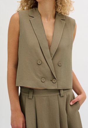 Kobieta ubrana w oliwkowo-zielony, bezrękawnikowy blezer dwurzędowy oraz pasujące, wysokie spodnie z ręką w kieszeni.