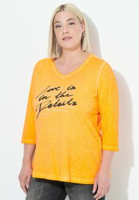Donna con taglio di capelli bob biondo, che indossa orecchini a cerchio, una maglietta arancione con scollatura a V e testo nero e bianco, e jeans scuri su sfondo bianco.