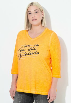 Frau mit blonden Bob-Haaren, die Ohrhänger trägt, ein oranges V-Ausschnitt-Shirt mit schwarz-weißer Schrift und dunklen Jeans vor weißem Hintergrund.