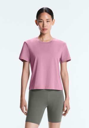 Kobieta stoi przodem, ubrana w różowy t-shirt z krótkim rękawem i szare dopasowane spodenki do kolan na jednolitym jasnym tle.