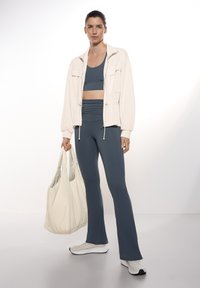 Veste bomber légère crème par-dessus un soutien-gorge de sport teal et un legging taille haute. Tient un sac fourre-tout beige doux, porte des chaussures de sport blanches.