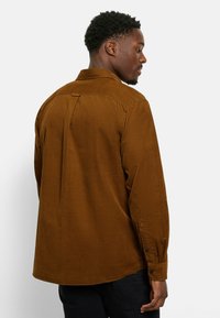 Braunes Cordhemd mit Rückenschlitz, spitzem Kragen und geknöpften Manschetten, das eine glatte Textur und eine lässige Passform aufweist.