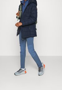 Veste matelassée bleu marine foncé avec capuche, couche zippée bleue, et pantalon bleu clair fuselé. Baskets grises et orange avec semelle texturée et lacets.