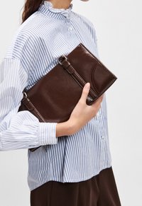 Personne portant un chemisier rayé bleu et blanc tenant une pochette en cuir marron foncé sur un fond clair uni.