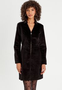 Robe en velours côtelé noir avec col et fermeture éclair à l'avant, manches longues et taille cintrée, associée à des collants à pois. Texture lisse.