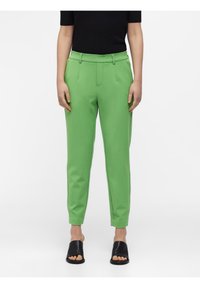Pantalon fuselé vert en tissu lisse avec taille haute, présentant des plis à l'avant et des poches latérales. Associé à des sandales noires à enfiler.