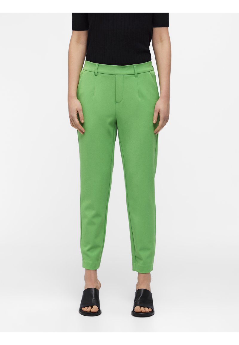 Pantalon fuselé vert en tissu lisse avec taille haute, présentant des plis à l'avant et des poches latérales. Associé à des sandales noires à enfiler.