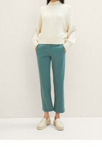 Crème ribbentrui met hoge hals, gecombineerd met teal cropped broeken. Schoeisel: crème loafers. Eenvoudig ontwerp, zachte texturen.