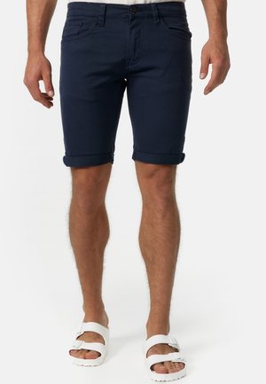 Homme portant un short bleu foncé retroussé et des sandales blanches à boucle, debout devant un fond uni.