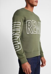 Felpa verde oliva con testo bianco in grassetto "REMIND" e design a righe sulle maniche. Presenta colletto girocollo e polsini a costine.