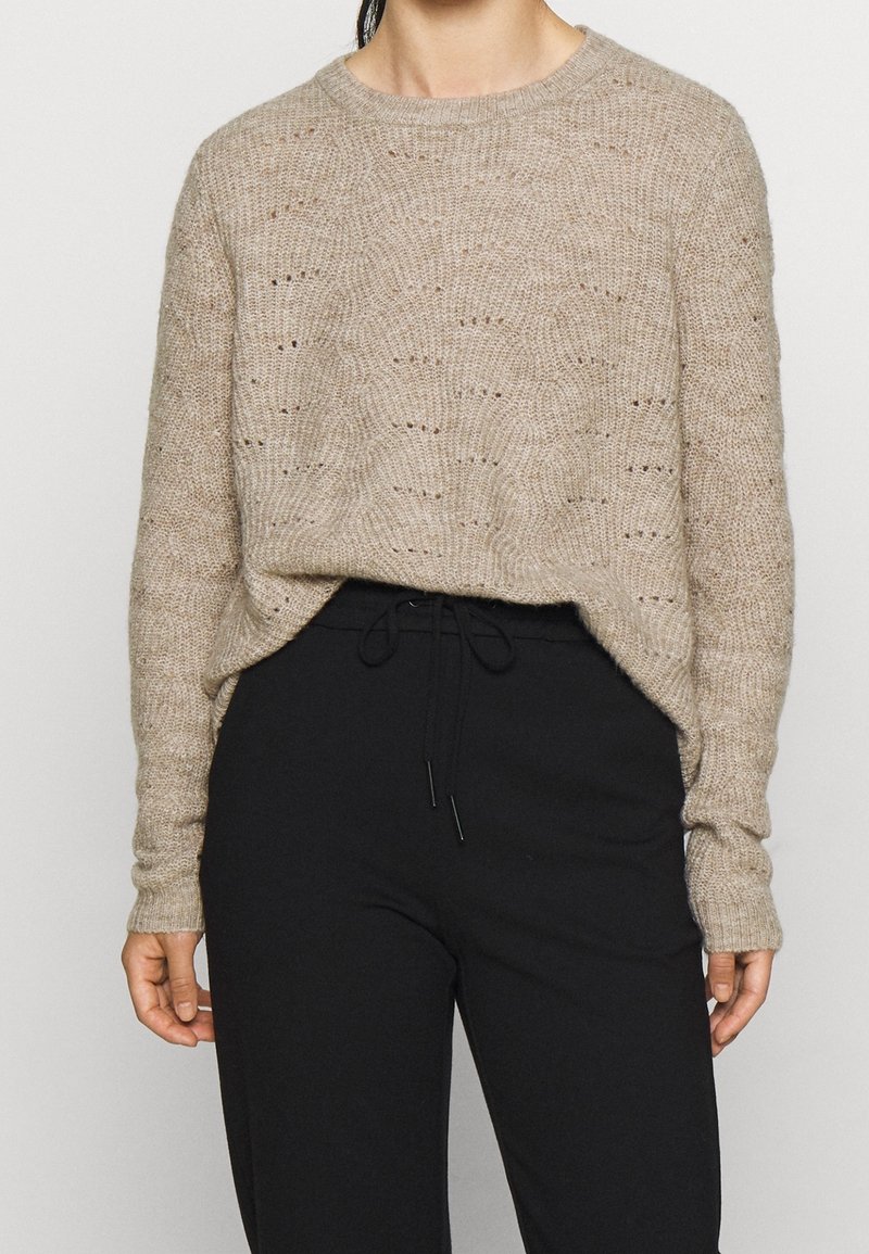 Pull beige en maille avec un col rond, présentant un motif ondulé texturé et des épaules tombantes, associé à un pantalon noir à taille élastique.