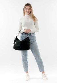 Schwarze Wildleder-Schultertasche mit silbernem Zubehör, die eine glatte Textur und eine geschwungene Form aufweist. Getragen von einem Model in heller Denim und weißen Sneakers.
