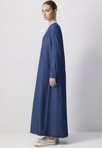 Robe maxi en denim bleu foncé avec des manches longues, coupe décontractée et fentes sur les côtés. Associée à des baskets beiges avec un ourlet effiloché.