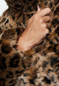 Cappotto in pelliccia sintetica con un motivo in stampa leopardata marrone e nera. Prima piano di una mano che afferra il morbido tessuto texturizzato lungo il colletto.