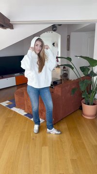 Sweatshirt branco com capuz, jeans azuis e sapatilhas prateadas. O fundo apresenta um sofá de bombazina castanha e uma planta verde. Pavimento em madeira.