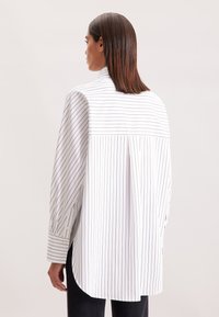 Vrouw met steil bruin haar die een losse witte blouse met dunne verticale en horizontale zwarte strepen draagt, gezien vanuit de rug.