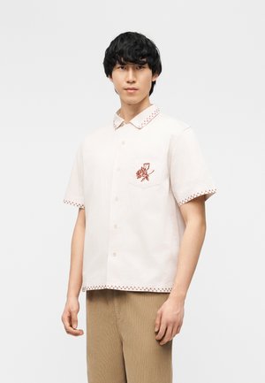 Noor mees, kes kannab lühikeste varrukatega valget nööbitavat särki, millel on taskul punane lilleline tikand ja punane õmblusääris, kombineeritud beežide pükstega.