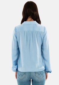Blusa azzurra chiara con colletto, dettagli plissettati sul retro e maniche lunghe. Abbinata a jeans blu aderenti con un design distintivo della tasca posteriore.