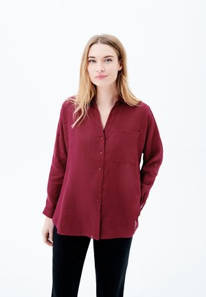 Camicia button-up bordeaux con colletto e tasca sul petto, realizzata in un tessuto leggero. Maniche lunghe con polsini abbottonati, indossata con pantaloni neri.