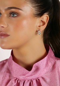 Silverörhängen med kedjelänkdesign och klara accenter. Bärs med en rosa, texturerad blus med hög krage. Huden ser slät ut.