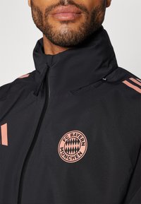 Musta vetoketjullinen takki, jossa on vaaleanpunainen FC Bayern München -logo. Ominaisuuksiin kuuluu korkea kaulus ja erottuvat raidat olkapäillä. Sileä pinta.