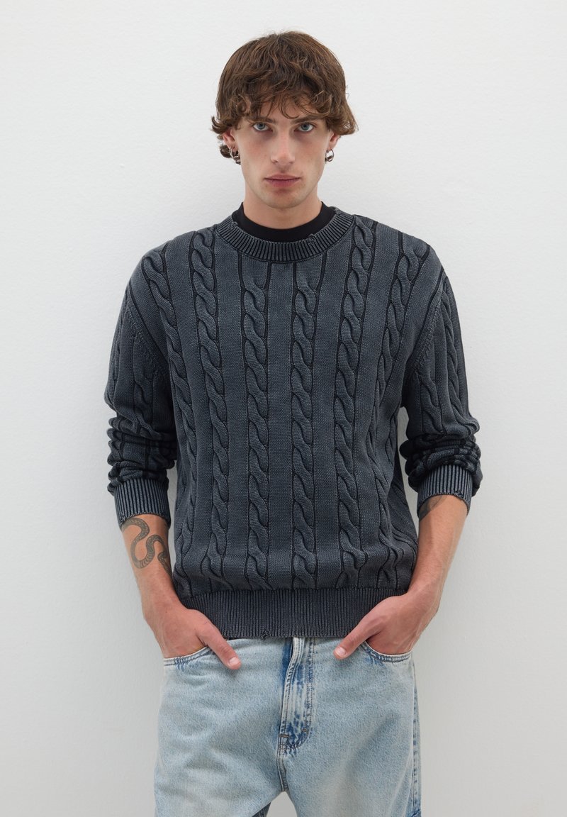 Maglione grigio scuro a trecce con colletto e polsini a coste, indossato sopra una maglia nera, abbinato a jeans in denim azzurro chiaro.