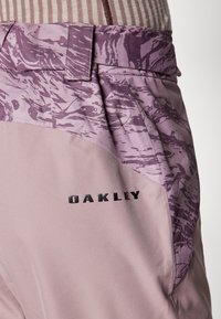Oakley-Hosen in einem helllila Farbton, mit einem seitlichen Panel in dunklerem lila mit abstraktem Muster und einem strukturierten Bund. Schwarzes Logodetail.