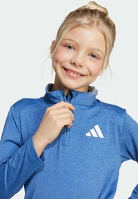 Zils sporta pulovers ar augstu apkakli, garām piedurknēm un ceturtdaļas rāvējslēdzēju; ar balto logo uz krūtīm un teksturētu audumu.