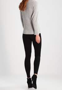 Žena oblečená v šedém žebrovaném svetru, černých legínách a černých vysokých podpatcích, stojící na bílém pozadí.