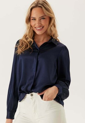 Marine satijnen blouse met lange mouwen en knoopmanchetten. Zachte textuur, met kraag ontwerp en subtiele zakdetails. Geko paired met witte jeans.