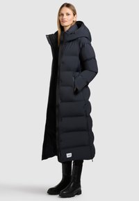 Langer schwarzer Daunenmantel mit hohem Kragen, großer Kapuze und gestepptem Muster. Verfügt über seitliche Taschen und ein Markenlabel nahe dem Saum.