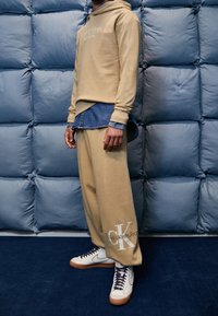 Beige hoodie met Calvin Klein logo, gedragen over eendenimshirt. Losse beige broek met CK-branding. Witte sneakers met oranje zolen.