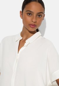 White short-sleeved shirt with a buttoned placket and soft collar. Smooth fabric with a relaxed fit, highlighting a clean, minimalist design.