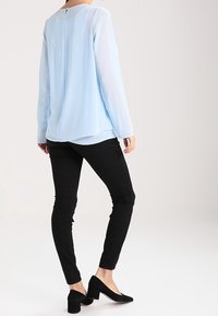Light blue long-sleeve blouse with a rounded neckline, sheer sleeves, and a back keyhole detail, paired with black form-fitting pants and black heels.