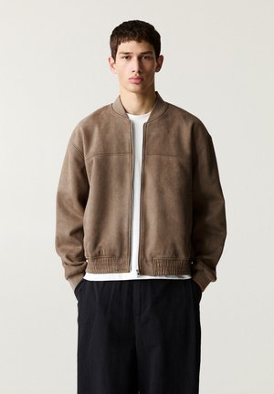 Brown suede bomber jacket with a zip closure, ribbed cuffs, and waistband. Worn over a white t-shirt, paired with black trousers.