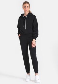 Czarna bluza z kapturem i kieszenią z przodu, w połączeniu z czarnymi spodniami dresowymi. Białe sznurówki i buty. Miękka tkanina i luźny krój.