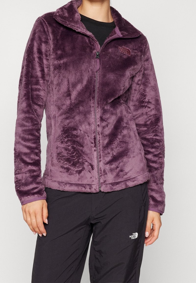 Person klädd i en plyschlila zip-up-jacka och svarta byxor med The North Face-logotyp, stående mot en enkel ljus bakgrund.