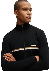 Schwarze Zip-Jacke mit hohem Kragen, ausgestattet mit horizontalen Streifen in Beige und Weiß. Auf der Vorderseite befindet sich der Schriftzug "BOSS" in weißen Buchstaben.