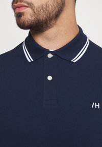 Námořnická polo trička s límečkem, zdobená bílými pruhy. Má dva knoflíky a vyšívané logo na hrudi.