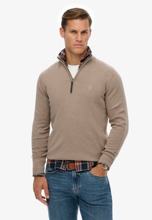 Mężczyzna w beżowym swetrze na suwak do połowy szyi, noszący pod nim czerwoną i niebieską koszulę w kratę, niebieskie dżinsy, brązowy pasek i zegarek na nadgarstku, stoi z jedną ręką w kieszeni.