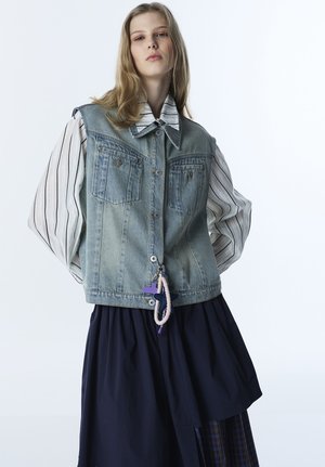 Junge Frau trägt ein gestreiftes Hemd mit Puffärmeln unter einer Jeansweste, kombiniert mit einem mehrlagigen marineblauen Rock und einem lilafarbenen Schlüsselanhänger.