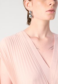 Light pink pleated dress with a V-neckline, paired with round metallic earrings. Smooth texture; delicate folds create visual interest.