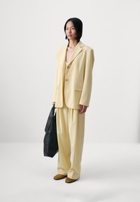 Yellow tailored blazer with single button and pocket, paired with wide-leg yellow trousers. Model holds a large black bag and wears brown shoes.