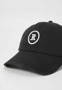 Casquette de baseball en coton noire avec une visière incurvée, présentant une lettre "R" brodée en blanc à l'intérieur d'un cercle sur le devant.
