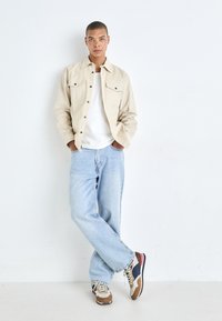 Beige button-up jacket over a white shirt, paired with light blue baggy jeans and multicolored sneakers. Textured fabric and relaxed fit.