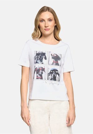 Femme blonde portant un T-shirt blanc illustré de quatre chiens noirs portant des lunettes de soleil à montures carrées et un pantalon beige clair.